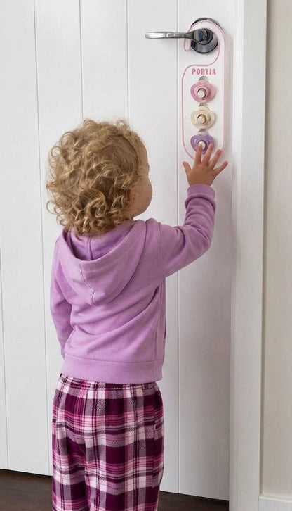 A toddler with curly blonde hair stands in front of a white door, pressing colorful buttons on a vertical door alarm device. The child is wearing a lavender hoodie and purple-and-white plaid pajama pants.